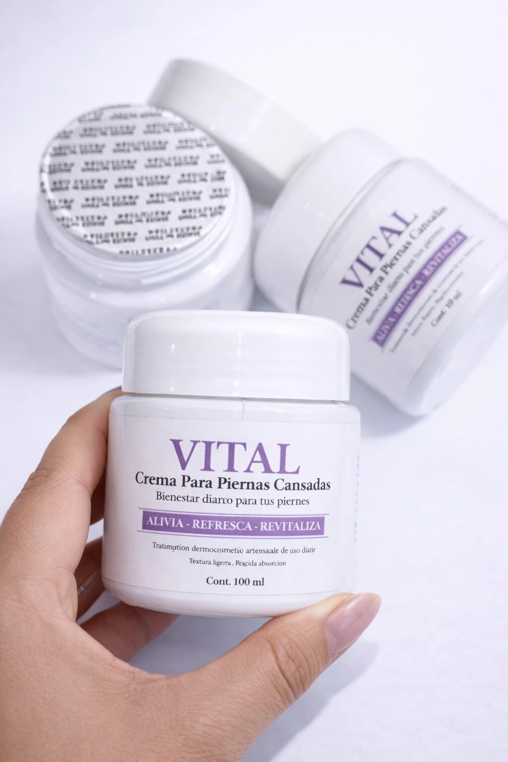 Vital Crema para Piernas Cansadas, presentación de 100 ml, con tapa blanca y etiqueta morada. Se muestra en mano junto a dos envases adicionales en fondo blanco.