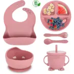 Set de vajilla para bebé en color rosa, incluye babero, plato dividido con frutas, cuchara, tenedor, taza con asa y bol con base antideslizante.