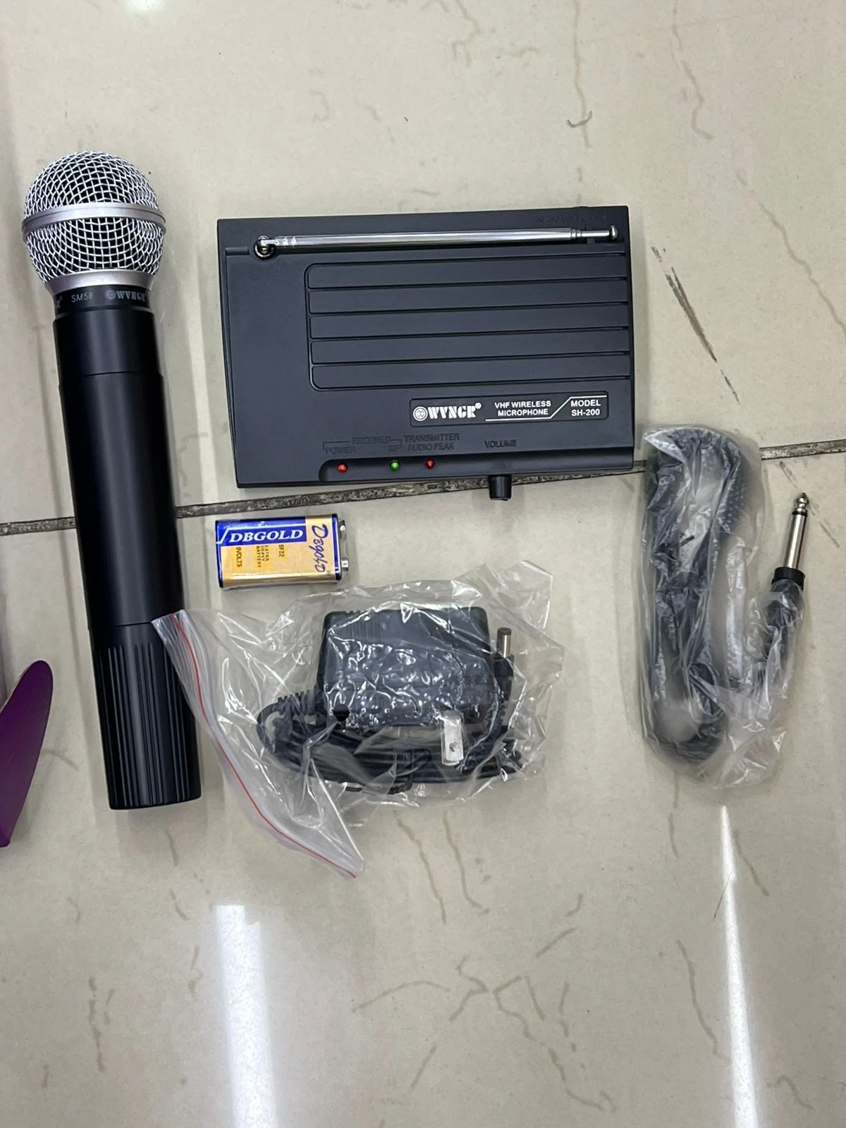Micrófono inalámbrico VHF Wyngir SH-200 con antena retráctil, base de carga, cable de audio, adaptador y pila, sobre superficie clara.