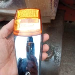 Indicador lateral compacto con carcasa cromada y lente ámbar, sostenido en mano para mostrar detalles de acabado y diseño.