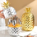 Set de cubiertos de acero inoxidable con estuche de piña en acabado dorado y plateado, mostrando utensilios organizados dentro del contenedor.