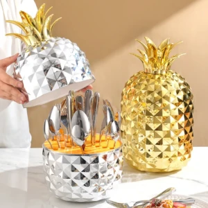 Set de cubiertos de acero inoxidable con estuche de piña en acabado dorado y plateado, mostrando utensilios organizados dentro del contenedor.