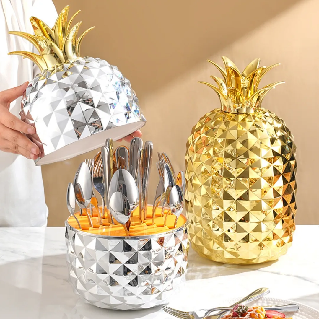 Set de cubiertos de acero inoxidable con estuche de piña en acabado dorado y plateado, mostrando utensilios organizados dentro del contenedor.