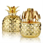Set de cubiertos de acero inoxidable con estuche de piña dorada, incluye tenedores, cuchillos y cucharas organizados en contenedor con forma de piña geométrica.