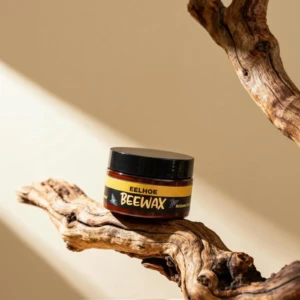 Cera de abeja natural en tarro negro con tapa, sobre rama de madera seca. Etiqueta amarilla con texto negro y blanco. Producto para restaurar brillo y proteger muebles y suelos.