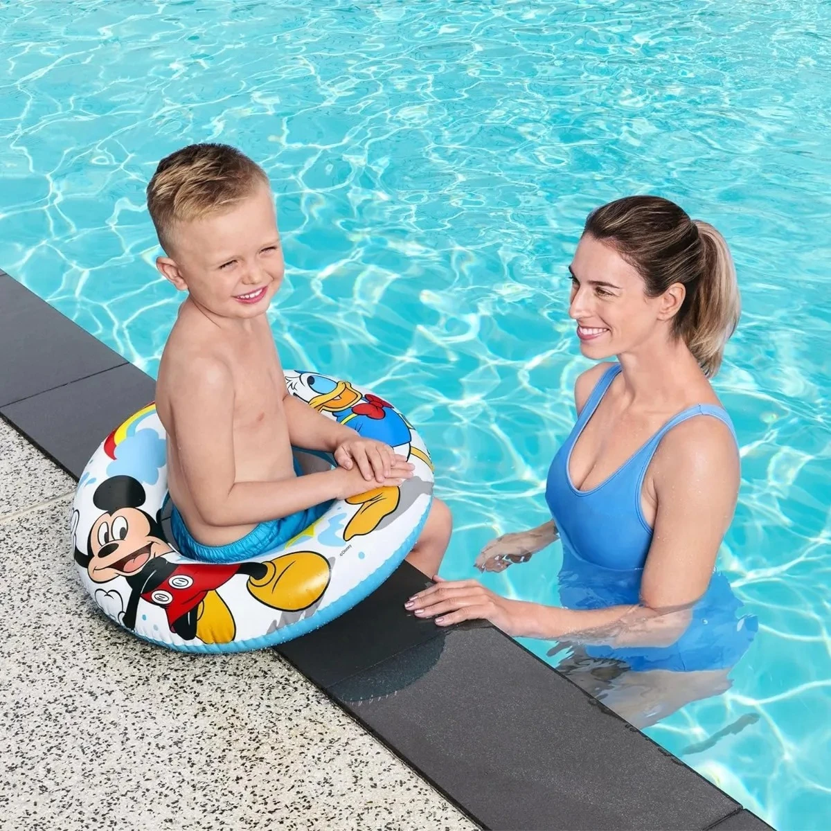 Niño usando flotador redondo de Mickey Mouse en piscina, con mujer adulta al lado. Flotador azul con personajes de Disney.