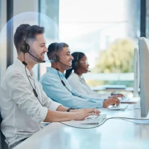 Tres operadores en call center usando diademas de audio con micrófono, sentados frente a computadoras en oficina moderna con luz natural.
