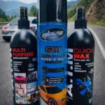 Kit de limpieza automotriz sin agua con tres botellas negras y un aerosol azul, mostrando etiquetas con nombres de productos y fotos de autos brillantes, sobre asfalto con carretera y montañas al fondo.