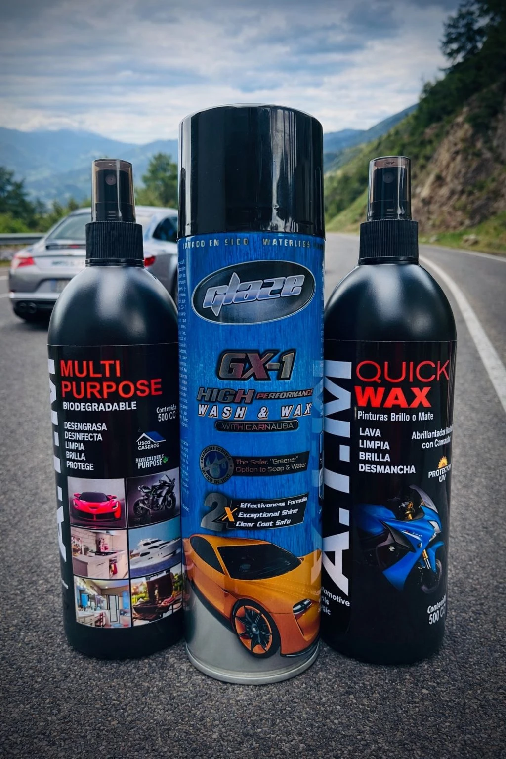 Kit de limpieza automotriz sin agua con tres botellas negras y un aerosol azul, mostrando etiquetas con nombres de productos y fotos de autos brillantes, sobre asfalto con carretera y montañas al fondo.