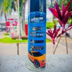 Spray de limpieza automotriz GX-1 en presentación azul con etiqueta que muestra auto naranja brillante, colocado sobre superficie gris con plantas de fondo.
