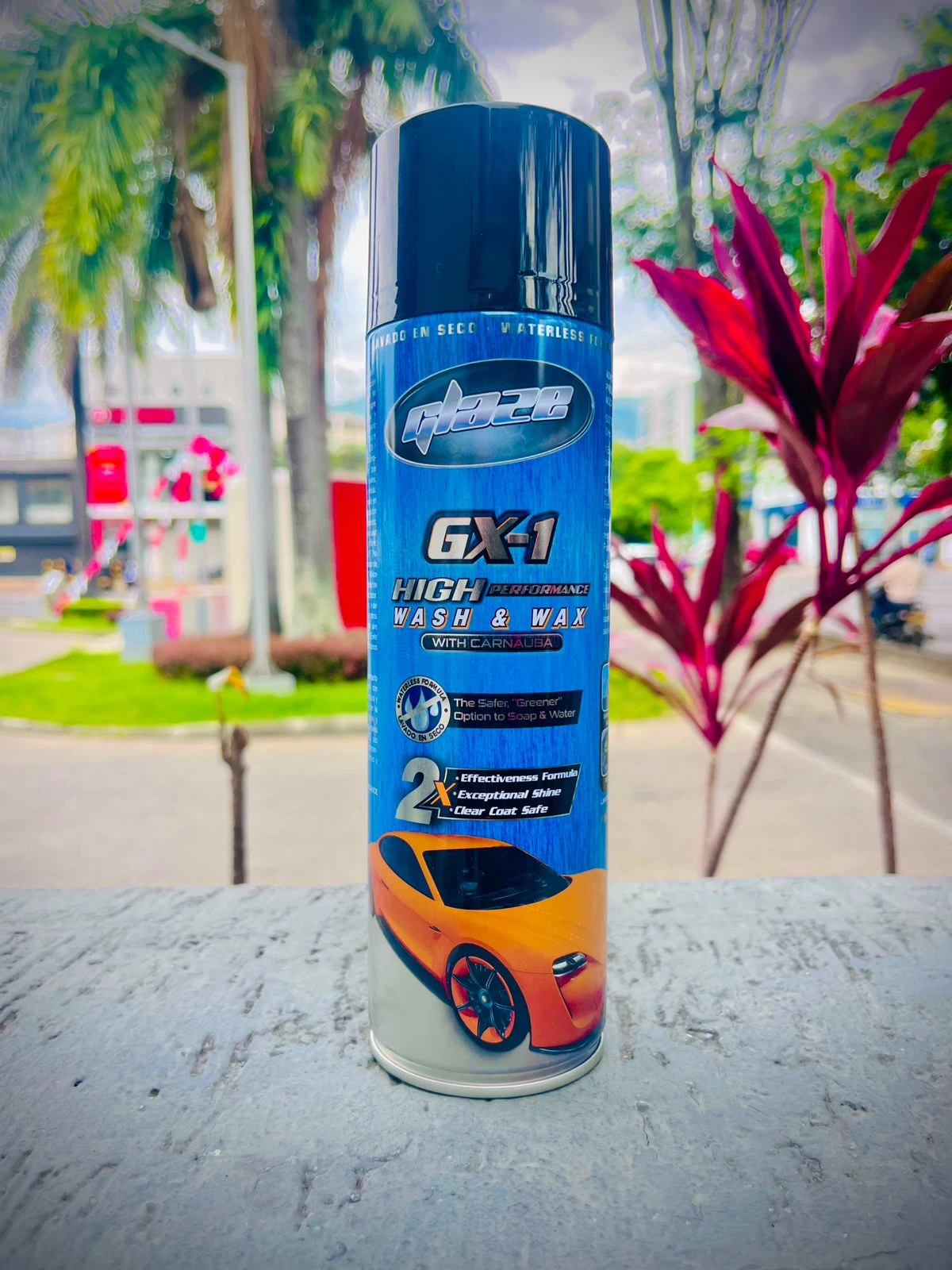 Spray de limpieza automotriz GX-1 en presentación azul con etiqueta que muestra auto naranja brillante, colocado sobre superficie gris con plantas de fondo.
