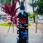 Botella negra de Quick Wax con boquilla spray, etiqueta que indica limpieza, brillo y desmancha, sobre superficie gris con plantas al fondo.