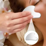 Gua sha de acero quirúrgico en uso sobre rostro, con lupa destacando su forma ergonómica y acabado pulido.