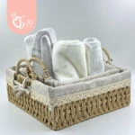Canasta tejida de ratan cuadrada con forro en tela beige y bordado, conteniendo toallas blancas enrolladas y accesorios de baño.