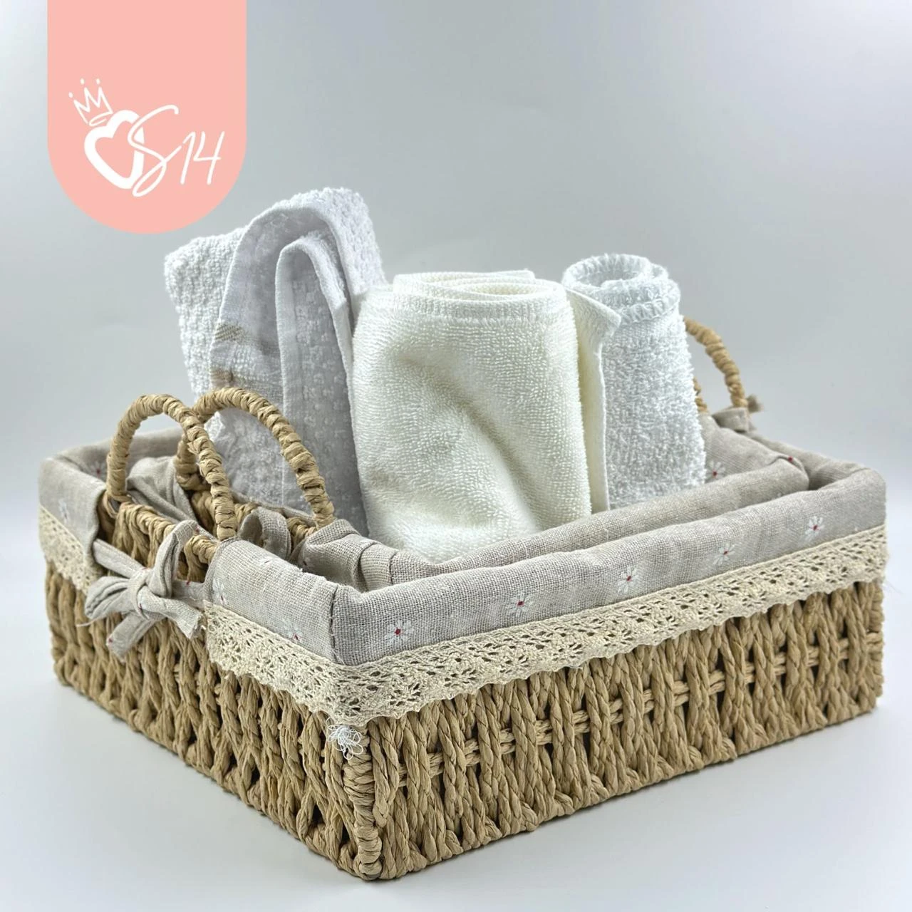 Canasta tejida de ratan cuadrada con forro en tela beige y bordado, conteniendo toallas blancas enrolladas y accesorios de baño.