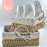 Canasta tejida de ratan cuadrada con forro interior, presentada en dos tamaños: una con toallas enrolladas y otra vacía. Incluye lazo decorativo y bordado floral. Ideal para organizar toallas en baños o salas.