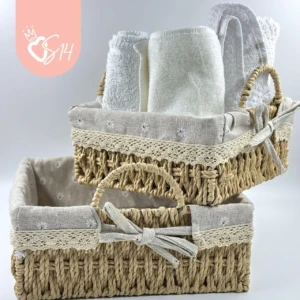 Canasta tejida de ratan cuadrada con forro interior, presentada en dos tamaños: una con toallas enrolladas y otra vacía. Incluye lazo decorativo y bordado floral. Ideal para organizar toallas en baños o salas.