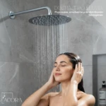 Regadera redonda de acero inoxidable 20x20 cm con flujo suave, instalada en baño de azulejos grises; mujer bajo la ducha con los ojos cerrados.