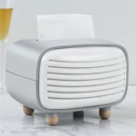 Dispensador de servilletas estilo radio retro en gris y blanco, con rejilla frontal y patas de madera, sobre superficie de mármol.