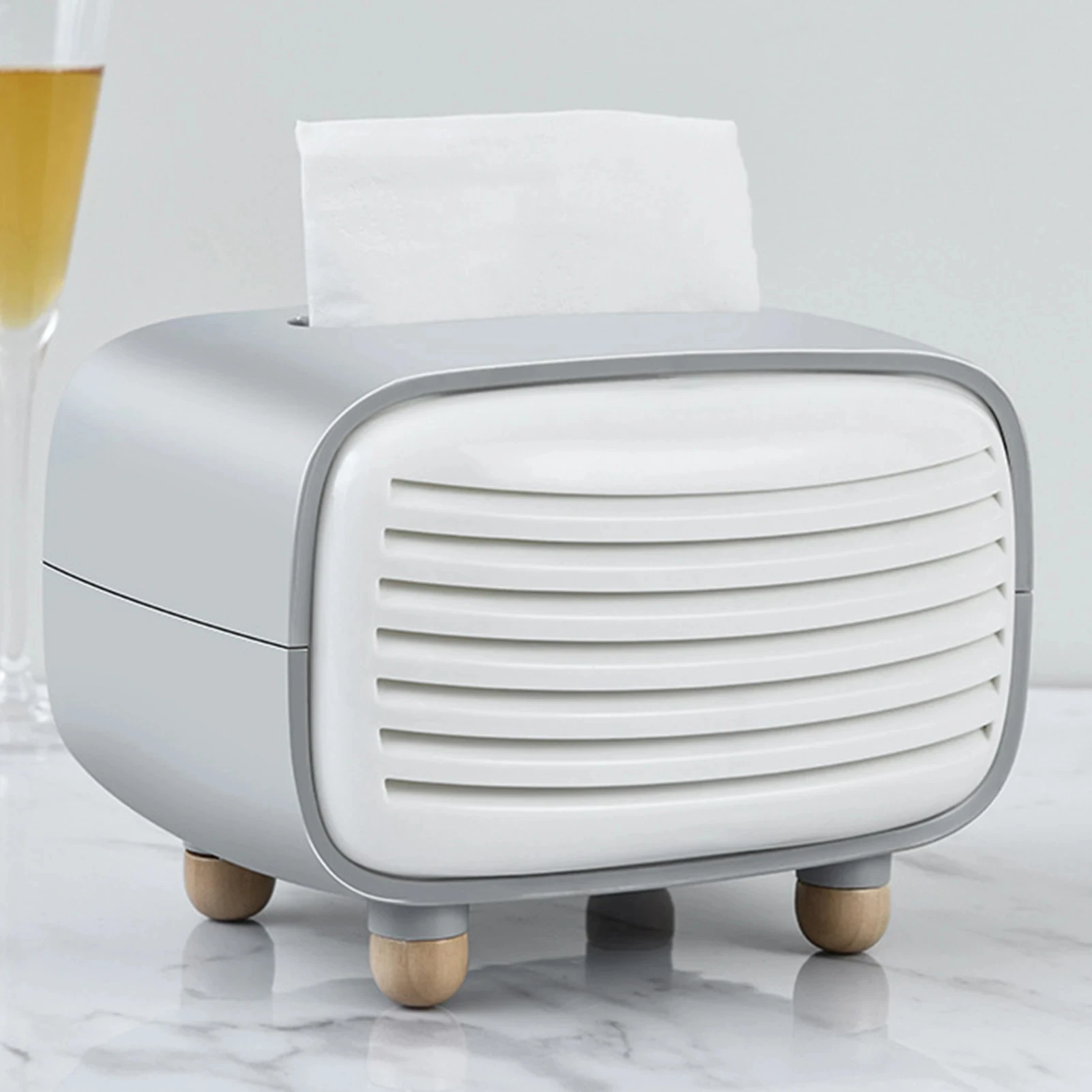 Dispensador de servilletas estilo radio retro en gris y blanco, con rejilla frontal y patas de madera, sobre superficie de mármol.