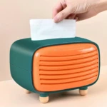 Dispensador de servilletas en forma de radio retro, color verde y naranja, con detalles en madera y una mano retirando una servilleta.