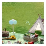 Alfombra picnic a cuadros verdes y blancos con cesta de mimbre, globos pastel, flores y accesorios de picnic sobre césped verde.