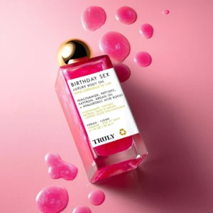 Botella de aceite corporal Truly Birthday Sex con líquido rosa brillante, etiqueta blanca con texto negro y tapa dorada, rodeada de gotas del mismo color sobre fondo rosa pálido.