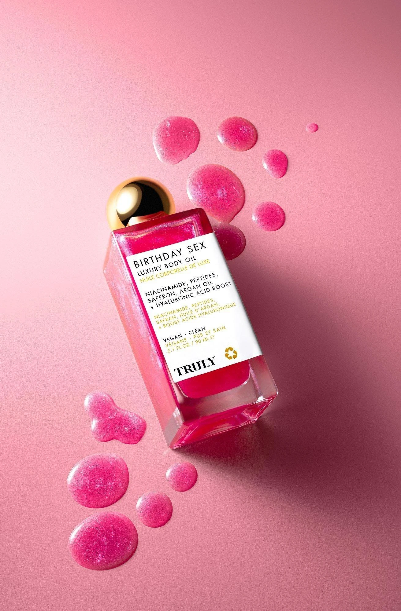 Botella de aceite corporal Truly Birthday Sex con líquido rosa brillante, etiqueta blanca con texto negro y tapa dorada, rodeada de gotas del mismo color sobre fondo rosa pálido.