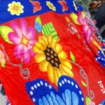 Cobija térmica roja con estampado de flores y mariposas en colores vivos, bordes azules y textura suave visible.