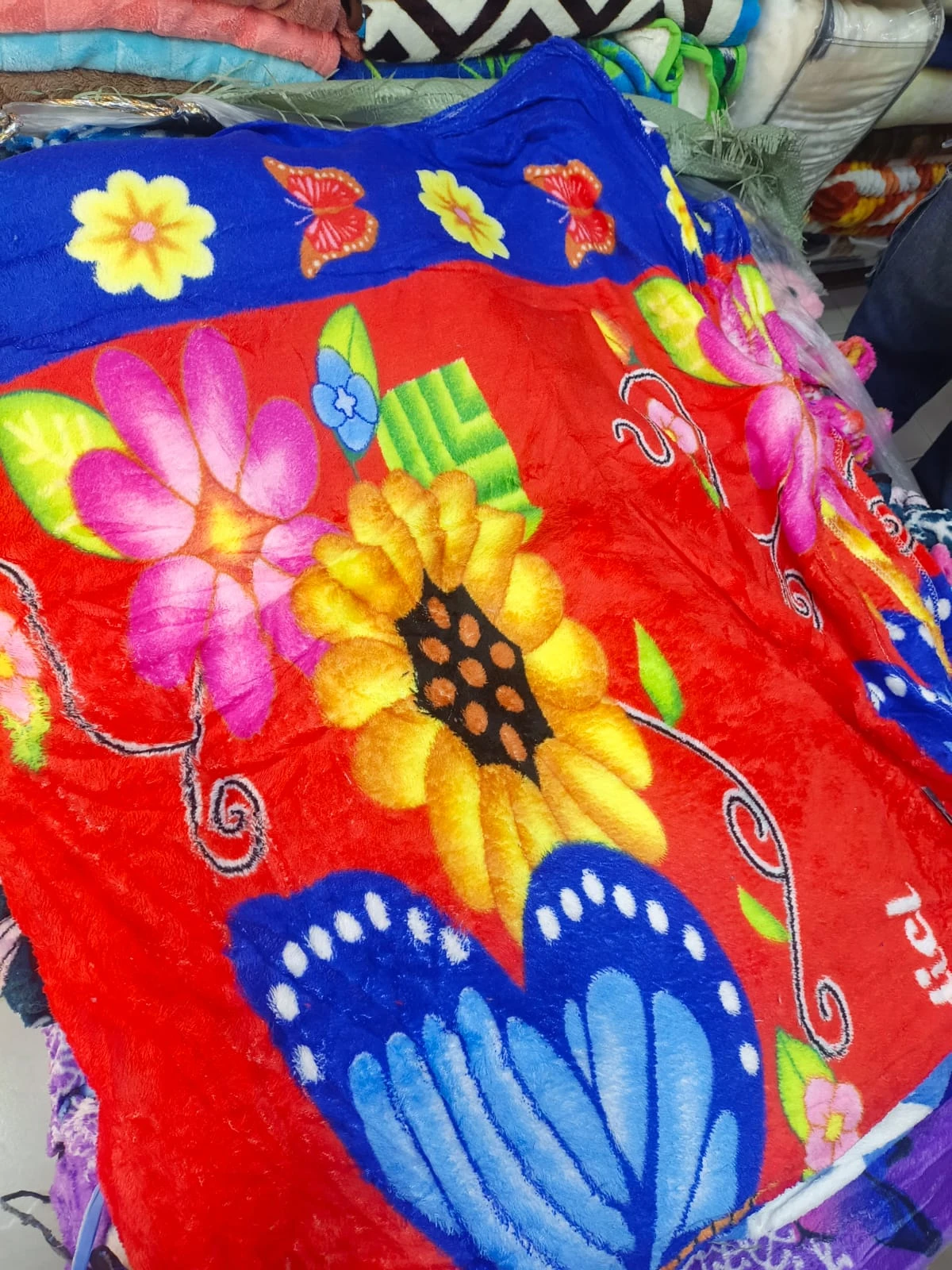 Cobija térmica roja con estampado de flores y mariposas en colores vivos, bordes azules y textura suave visible.