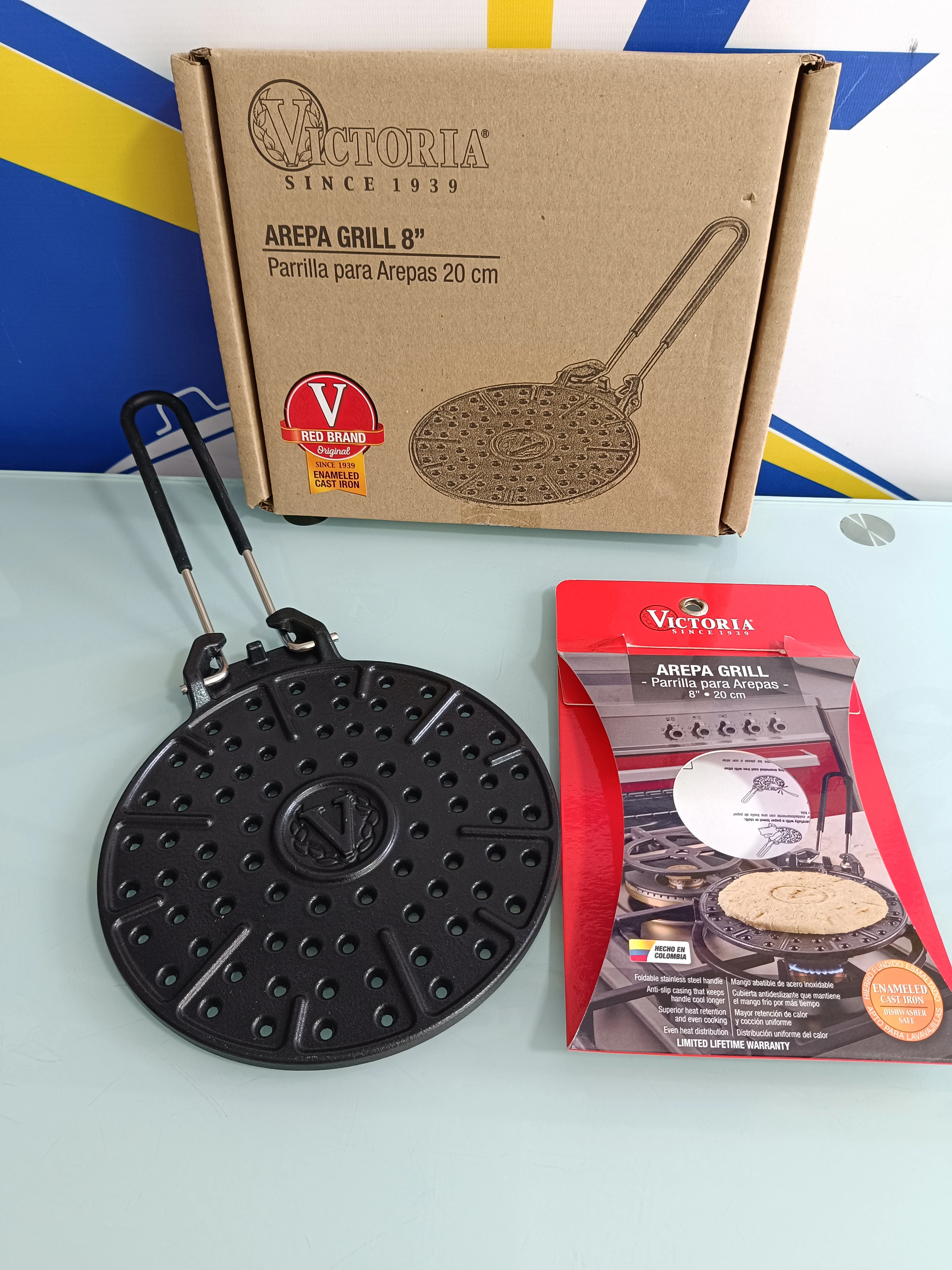 Parrilla para arepas de hierro fundido, color negro, con mango y caja original Victoria, junto a su empaque rojo.