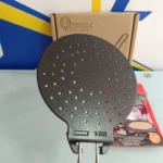 Parrilla arepas Victoria de hierro fundido con perforaciones circulares, mango metálico y caja de cartón al fondo.