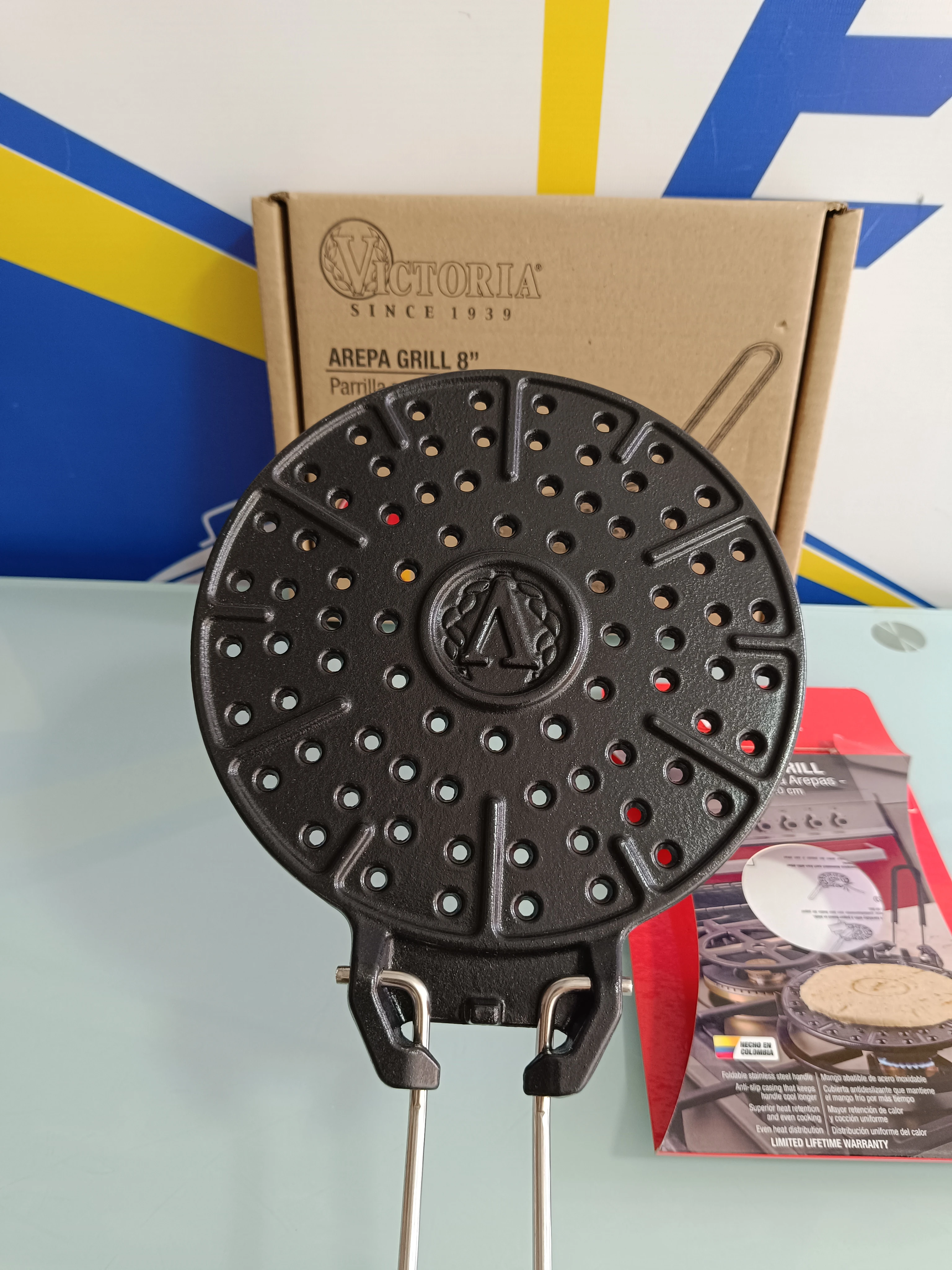 Parrilla arepas Victoria de hierro fundido con superficie perforada y empuñaduras metálicas, mostrada sobre su caja original.