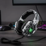 Diadema gamer Onikuma K8 en camuflaje blanco con micrófono y luces LED verdes, sobre escritorio con teclado y mouse.