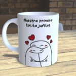 Taza de cerámica blanca con diseño de dos figuras abrazadas y corazones rojos, texto que dice "Nuestra primera tacita juntos", sobre mesa de madera.
