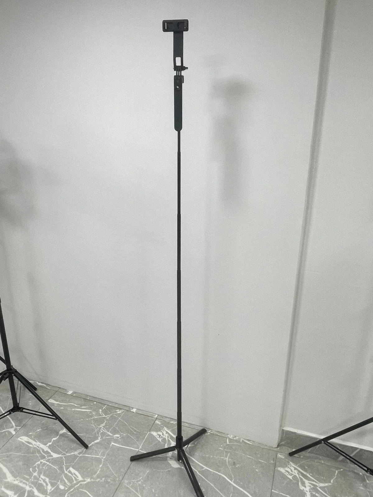 Trípode P205 negro con brazo extensible y base de tres patas, montado sobre piso de baldosas blancas con vetas grises.