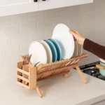 Organizador de vajilla en bambú de doble nivel con platos blancos y azules, cubiertos y utensilios en compartimento lateral, sobre encimera blanca.