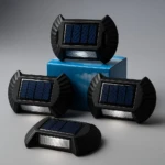 Cuatro lámparas solares negras con panel fotovoltaico y luz LED frontal, dispuestas sobre caja azul con cielo estrellado.