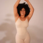 Faja moldeadora color crema, espalda descubierta, define cintura y mejora postura. Modelo con cabello afro, brazos levantados cruzados.