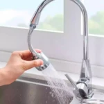 Llave de cocina con extensión flexible en uso, con mano ajustando la boquilla. Agua fluyendo hacia el fregadero. Diseño cromado, instalación visible junto a ventana.