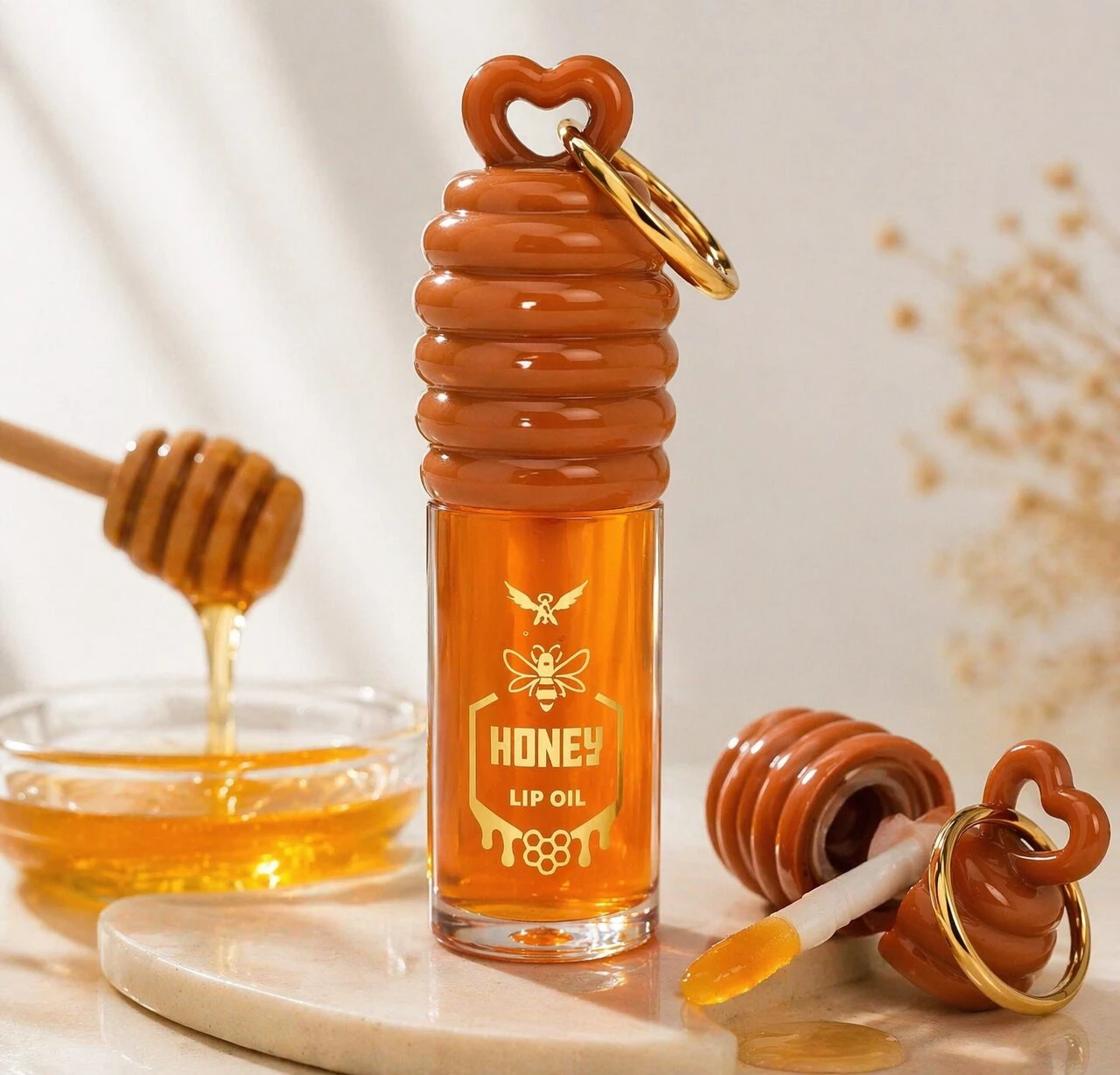 Aceite labial en frasco de vidrio con diseño de panal de miel, tapa en forma de corazón y anillo metálico, acompañado de abeja dorada y etiqueta que dice "Honey Lip Oil".