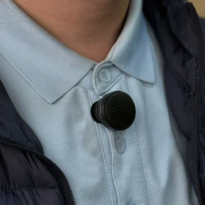 Micrófono solapa inalámbrico negro con batería integrada, colocado en la solapa de camisa azul claro, ideal para grabación profesional móvil.