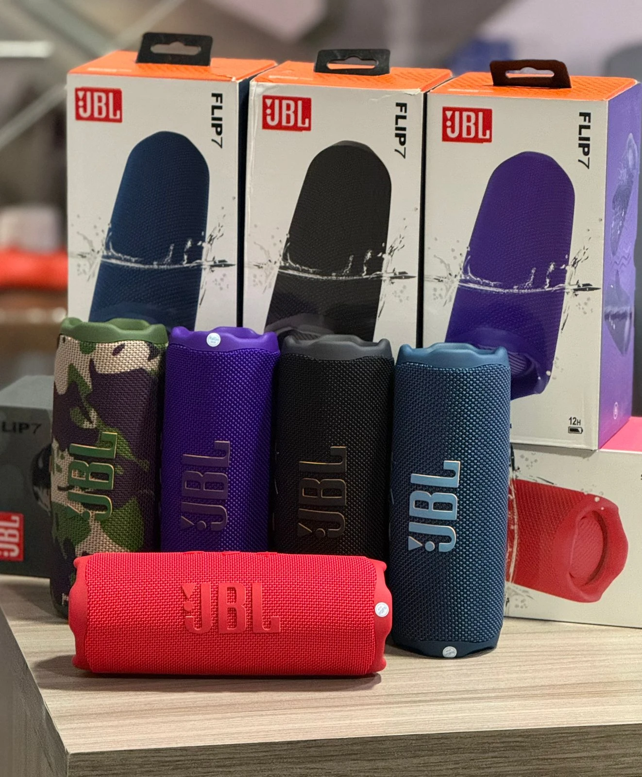 Cuatro parlantes JBL Flip 7 en colores azul, negro, morado y rojo, con caja de presentación naranja y morada al fondo.