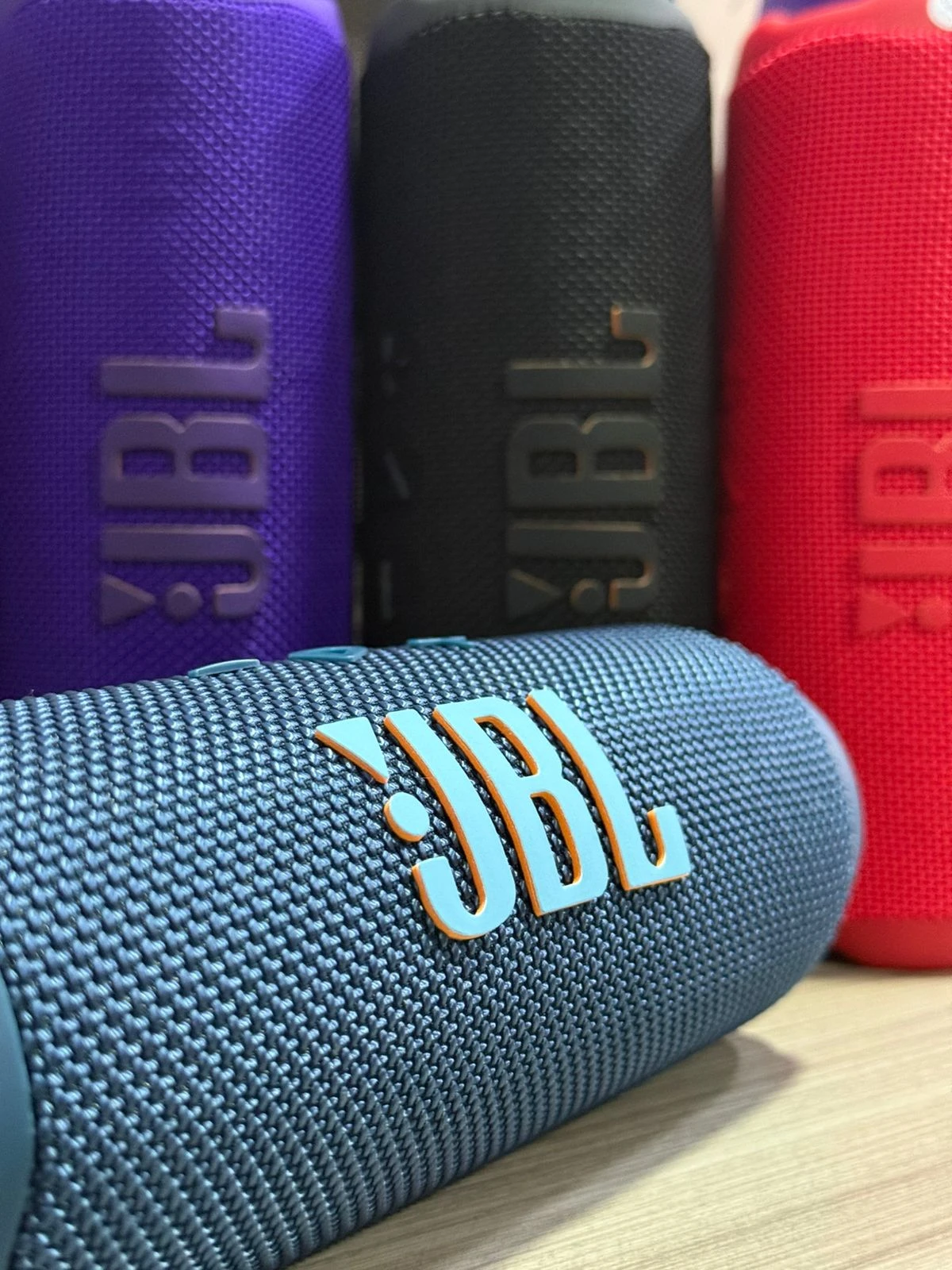 Cuatro parlantes JBL Flip 7 en colores azul, negro, rojo y morado, con textura de malla y logo visible, dispuestos sobre superficie de madera.