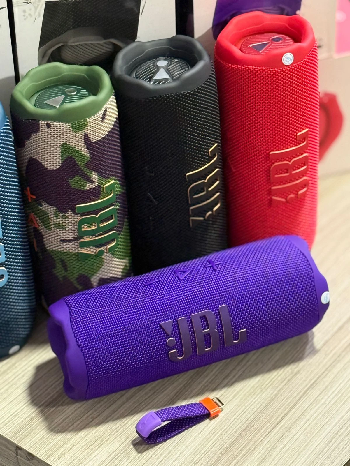 Cuatro parlantes JBL Flip 7 en colores verde camuflaje, negro, rojo y morado, con textura de malla y logotipo visible, dispuestos sobre superficie de madera.