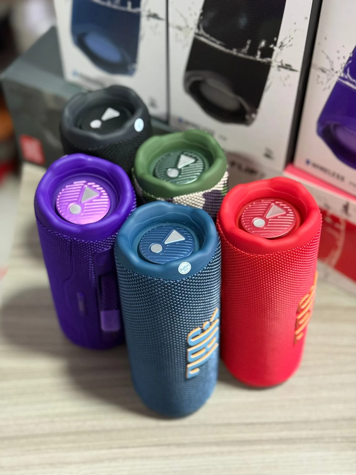 Cinco parlantes JBL Flip 7 en colores morado, azul, rojo, verde y negro, dispuestos sobre una superficie clara con cajas de empaque al fondo.