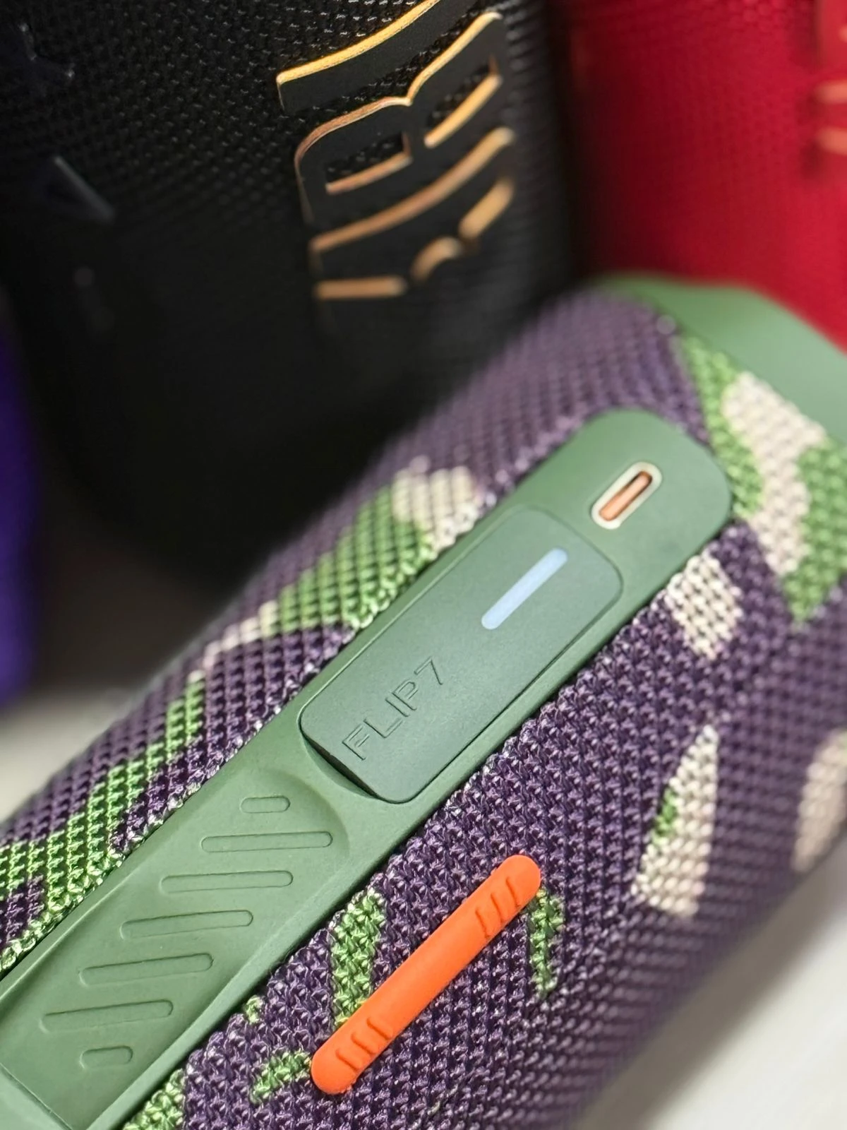Parlante portátil JBL Flip 7 en color verde con patrón geométrico, mostrando controles táctiles y botón de encendido naranja.