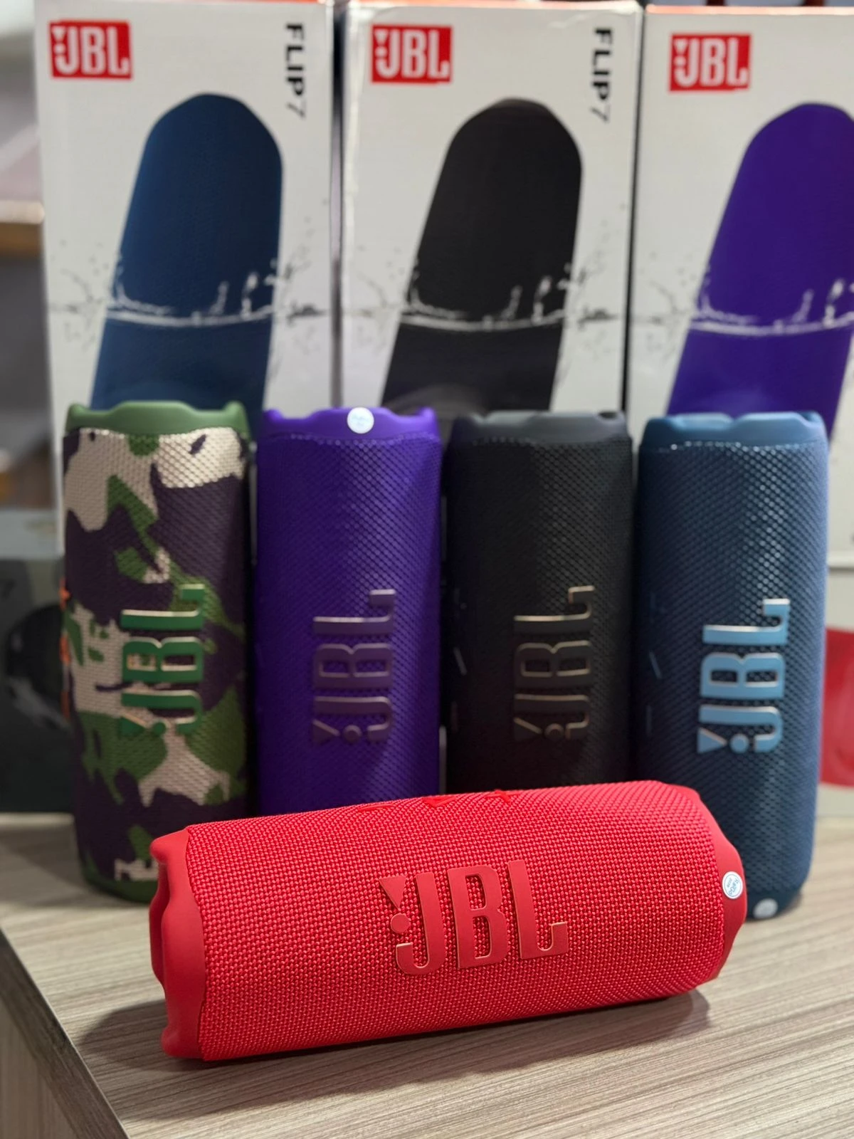 Cuatro parlantes JBL Flip 7 en colores verde camuflaje, morado, negro y azul, con caja de venta al por menor detrás, y uno rojo en primer plano sobre superficie de madera.