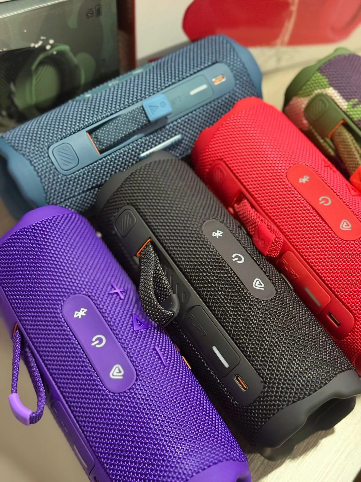 Cuatro parlantes portátiles JBL Flip 7 en colores azul, rojo, negro y morado, con textura de malla y controles táctiles visibles.