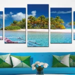 Reloj políptico de cinco paneles con paisaje marino tropical, cielo azul, palmeras y botes en aguas turquesas, sobre sofá azul con cojines verdes.